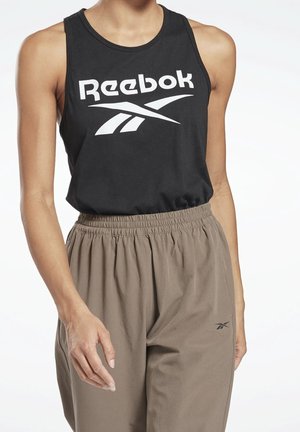 Kvinde iført en sort Reebok ærmeløs top og løse taupebukser med elastik i taljen, stående foran en hvid baggrund.