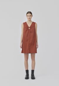 Robe sans manches couleur rouille avec un décolleté en V et un détail de nœud au centre. Fabriquée en tissu léger, elle a une silhouette ample en forme de A.