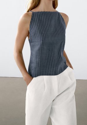 Haut sans manches bleu marine à fines rayures associé à un pantalon blanc ample, mains dans les poches, fond neutre.