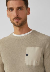 Beżowy sweter z prążkowanym wzorem, okrągłym dekoltem i małą kieszonką. Kieszonka ma ciemny guzik oraz detal z logo. Miękka tekstura.