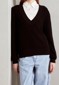 Schwarzer, gerippter Strickpullover mit tiefem V-Ausschnitt, langen Ärmeln und lockerer Passform. Getragen über einem weißen Hemd mit Kragen und kombiniert mit hellblauen Jeans.