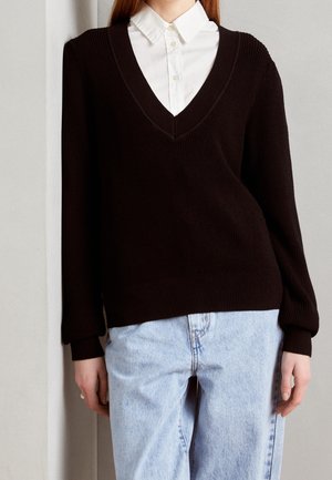 Pull en tricot noir côtelé avec un décolleté en V profond, des manches longues et une coupe décontractée. Porté sur une chemise blanche à col et associé à un jean bleu clair.