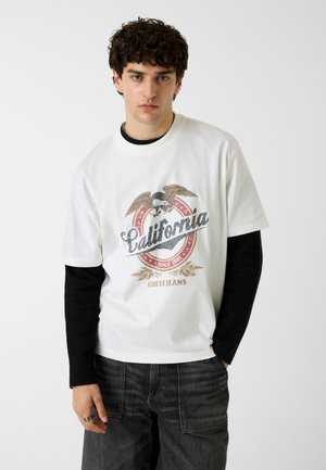 Young man with curly hair wears layered shirts, white over black, featuring a “California Guess Jeans” graphic with an eagle. Neutral background.