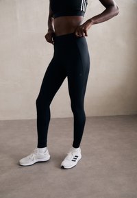 Fekete, magas derekú leggings, amelyen a combon logó található; fehér, fekete kiegészítőkkel ellátott tornacipővel párosítva. Sima, rugalmas anyag.