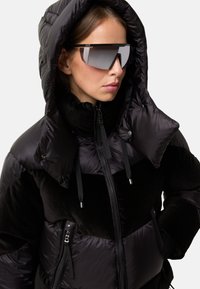 Puffetjakke i sort og struktureret sort stof med hætte, høj krave, lynlås lukning og reflekterende solbriller.