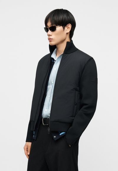 Άνδρας με μαύρο μπουφάν πάνω από μπλε ριγέ πουκάμισο, μαύρο παντελόνι, γυαλιά ηλίου, στέκεται με το ένα χέρι στην τσέπη μπροστά σε απλό φόντο.