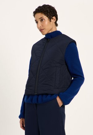 Junge Person trägt eine ärmellose gesteppte marineblaue Weste über einem blauen Rollkragenpullover und dunklen Hosen, mit einer Hand in der Tasche.