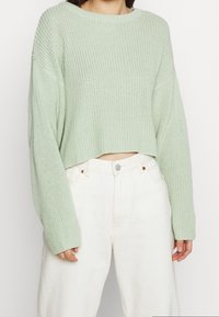 Vaaleanvihreä neulottu lyhyt villapaita, jossa leveä pääntie ja ylikokoiset hihat, yhdistetty valkoisiin korkeavyötäröisiin housuihin. Näkyvissä kankaan tekstuuri.