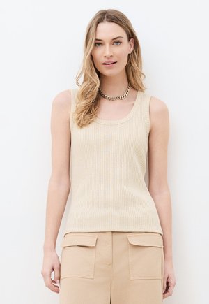 Vaaleaverikköinen nainen, joka pukeutunut beigeen ribbineulottuun hihattomaan toppiin, beigeihin cargohousuihin ja kultaketjuun, seisoo valkoista taustaa vasten.