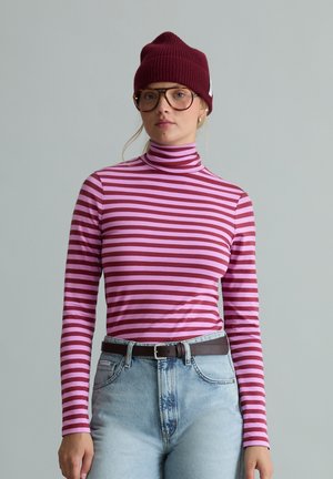 Frau trägt einen rot-rosa gestreiften Rollkragenpullover, hellblaue Jeans mit schwarzem Gürtel, große Brille und eine dunkelrote gestrickte Mütze.