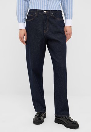 Person wearing dark blue straight-leg jeans, a blue and white striped shirt, and black chunky-soled loafers standing on a white background.