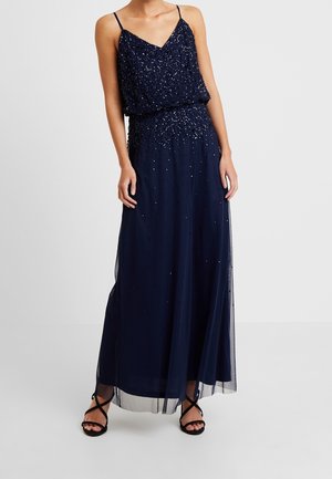 Kvinde iført en navyblå maxi-kjole uden ærmer med palietdekorationer og sorte højhælede sandaler med stropper, stående foran en ensfarvet baggrund.