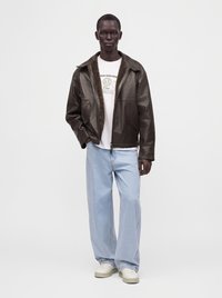Mann trägt eine dunkelbraune Lederjacke über einem weißen bedruckten T-Shirt, hellblaue Jeans mit weitem Bein und weiße Sneakers, steht vor einem einfarbigen Hintergrund.