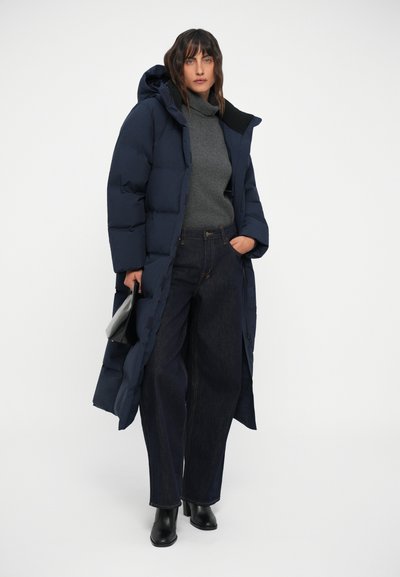Mornarska duga puffer jakna s kapuljačom, sivi turtleneck, široke tamne traperice i crne čizme do gležnjeva. Glatke teksture i minimalni detalji.