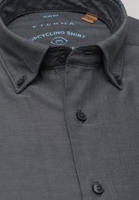Chemise upcyclée coupe ajustée en tissu gris foncé. Elle présente un col boutonné, six boutons noirs et un motif texturé. Étiquette Eterna à l'intérieur.