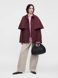 Personne portant une veste oversize bordeaux et un pantalon large beige clair, tenant un sac à main noir avec des poignées rondes.