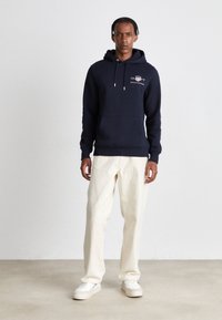 Mörkblå hoodie med framficka och dragsko på huvan, kombinerad med krämfärgade vida byxor och vita sneakers. Slät tygtextur.