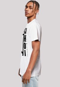 White cotton t-shirt with short sleeves, featuring bold black text and graphic design on the left side. Casual fit and crew neck style.