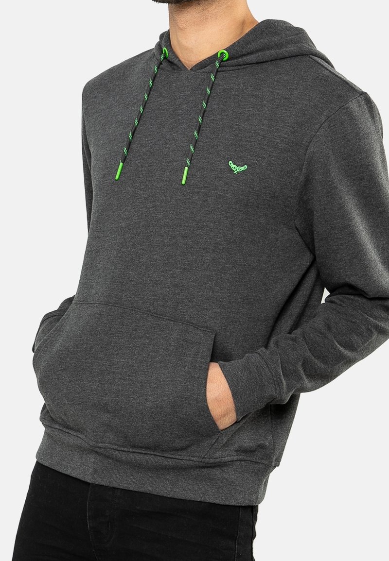 Homme portant un sweat à capuche gris foncé avec des cordons verts et un petit logo vert, les mains dans la poche avant, sur fond blanc.