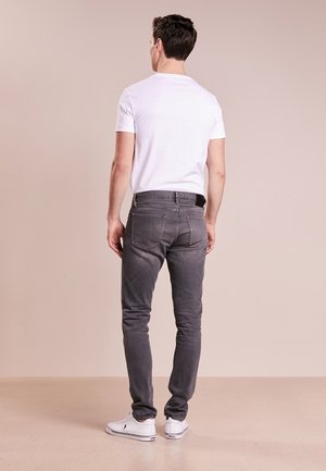 En man som står med ryggen vänd, iklädd en vit t-shirt, grå slim-fit jeans och vita sneakers mot en enfärgad beige bakgrund.