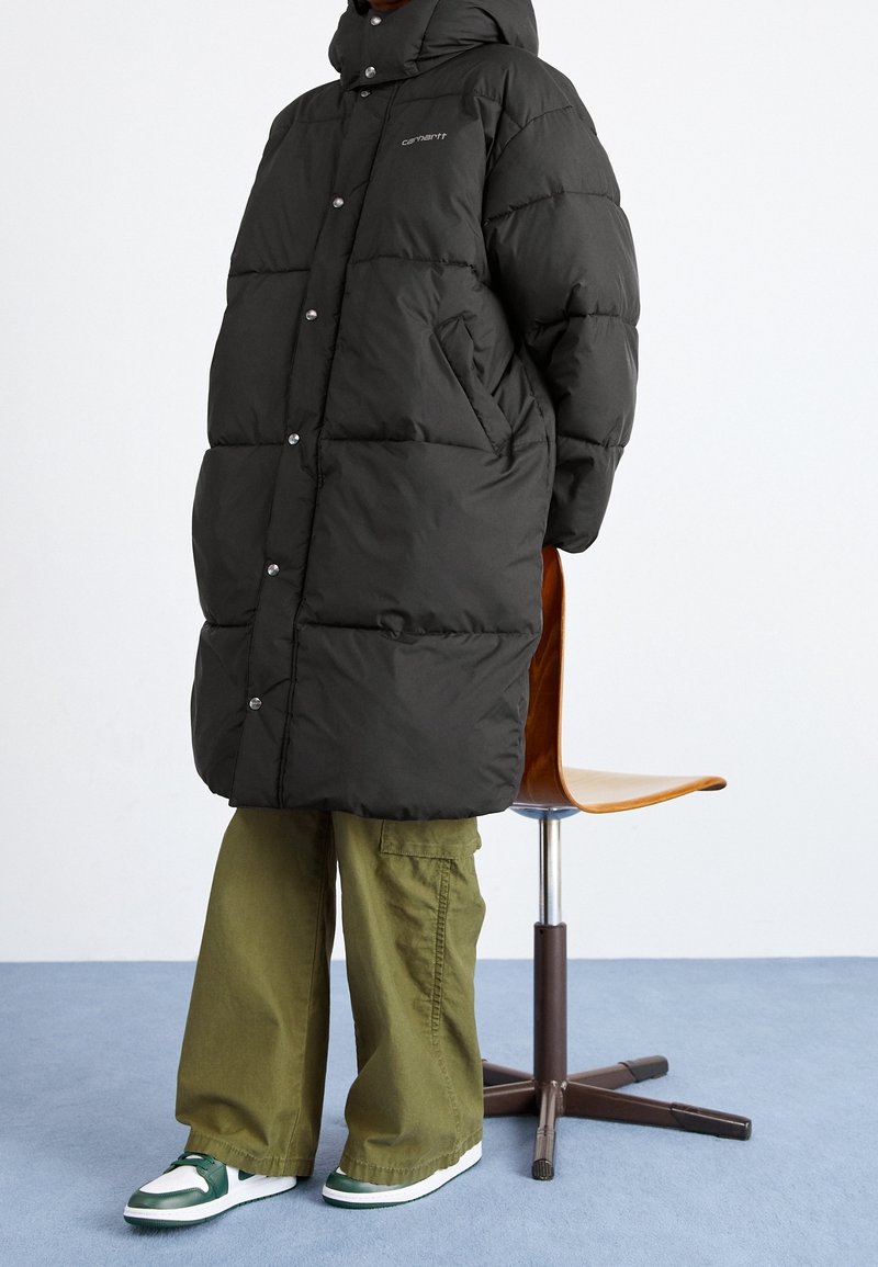 Zwarte pufferjas met capuchon en drukknoopsluiting, gecombineerd met olijfgroene cargobroek en sneakers met groene accenten. Op de achtergrond staat een stoel.