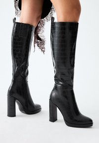 Botas negras hasta la rodilla hechas de cuero sintético texturizado con un patrón de cocodrilo, que presentan un tacón grueso y una cremallera lateral para el cierre.
