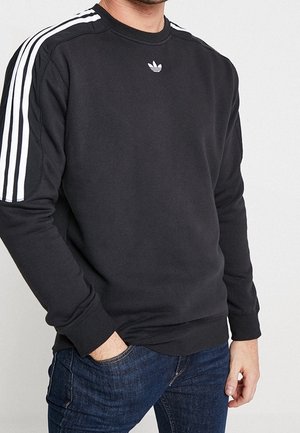 Mies, jolla on musta Adidas-collegepaita valkoisilla hihojen raidoilla ja rinnassa logolla, yhdistettynä tummansinisiin farkkuihin.