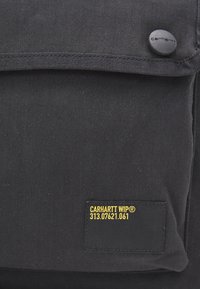Černá látková taška s klopou a zapínáním na knoflík, s černou nášivkou se žlutým textem "CARHARTT WIP® 313.07621.061".