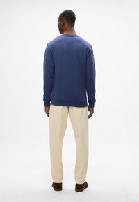 Mornarsko modra sweatshirt s okroglim ovratnikom, z ribastimi manšetami in robom, oblečena z svetlo bež hlačami in rjavimi čevlji, pogled od zadaj.