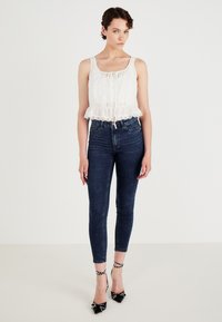 Witte crop top met kantafwerking en een trekkoord bij de hals, gecombineerd met donkerblauwe skinny jeans. Zwarte hakken met studded accenten.