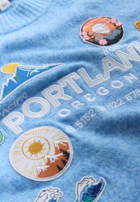 Tissu bleu avec des écussons brodés comprenant des montagnes, le soleil, des vagues et le texte "PORTLAND OREGON" avec des coordonnées géographiques.
