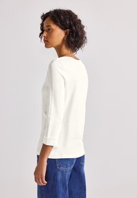 Haut blanc à manches longues avec un col rond, porté avec un jean en denim bleu. Texture lisse, design simple et manches ajustées se terminant au poignet.