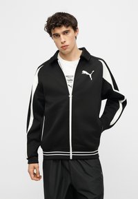 Νεαρός άνδρας που φοράει μαύρο-λευκό μπουφάν Puma με φερμουάρ και μαύρο παντελόνι, στέκεται μπροστά σε απλό φωτεινό φόντο.