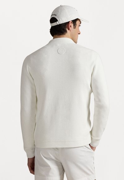 Pull en tricot blanc avec une texture côtelée, col rond et logo dans le dos, associé à un short blanc et une casquette blanche.