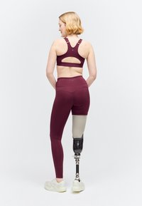 Jeune femme avec une prothèse de jambe sous le genou, portant un soutien-gorge de sport bordeaux, des leggings et des baskets blanches, de dos sur un fond blanc.