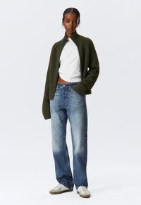 Cardigan de punto verde con cremallera, camiseta blanca, jeans de mezclilla azules y zapatillas beige. Tejido texturizado con puños acanalados y un corte corto.