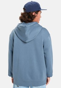 Haut à capuche bleu en mélange de coton, doté d'une grande capuche, de manches longues et d'une coupe décontractée. Finition avec un petit logo en détail au bas.