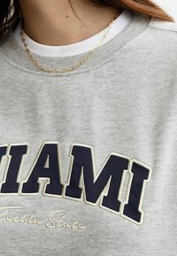 Grijze sweatshirt met "MIAMI" in grote marineblauwe letters, met "Florida State" in kleinere cursieve letters eronder. Draagt een witte T-shirt eronder.