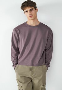 Langärmliges T-Shirt mit Rundhalsausschnitt in gedämpftem Mauve, aus weichem Baumwollstoff gefertigt. Kombiniert mit lockeren, khakifarbenen Cargohosen mit seitlichen Taschen.