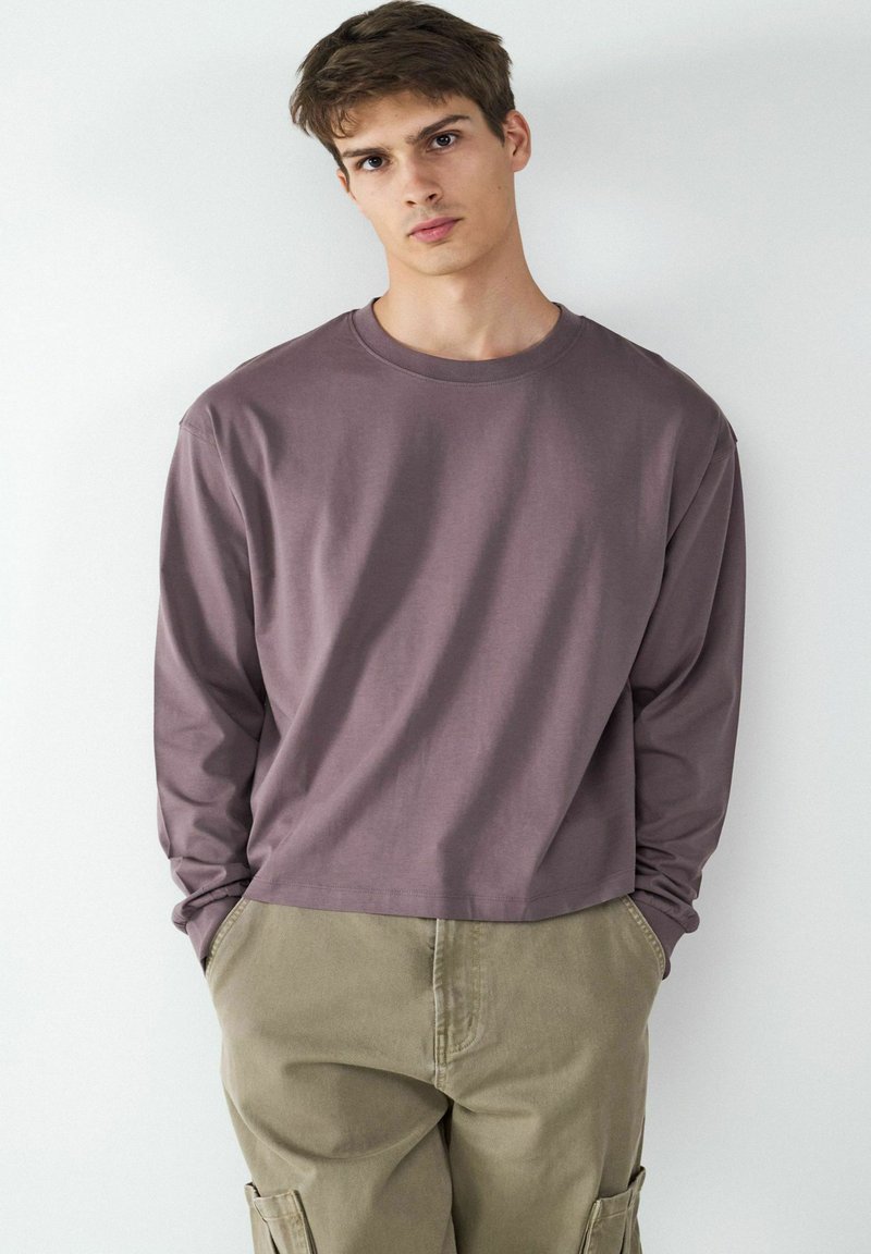 Langärmliges T-Shirt mit Rundhalsausschnitt in gedämpftem Mauve, aus weichem Baumwollstoff gefertigt. Kombiniert mit lockeren, khakifarbenen Cargohosen mit seitlichen Taschen.