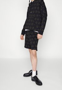 Black hoodie and shorts featuring a yellow smiley face pattern. The outfit has a relaxed fit, with textured fabric and ribbed cuffs. Black shoes complete the look.