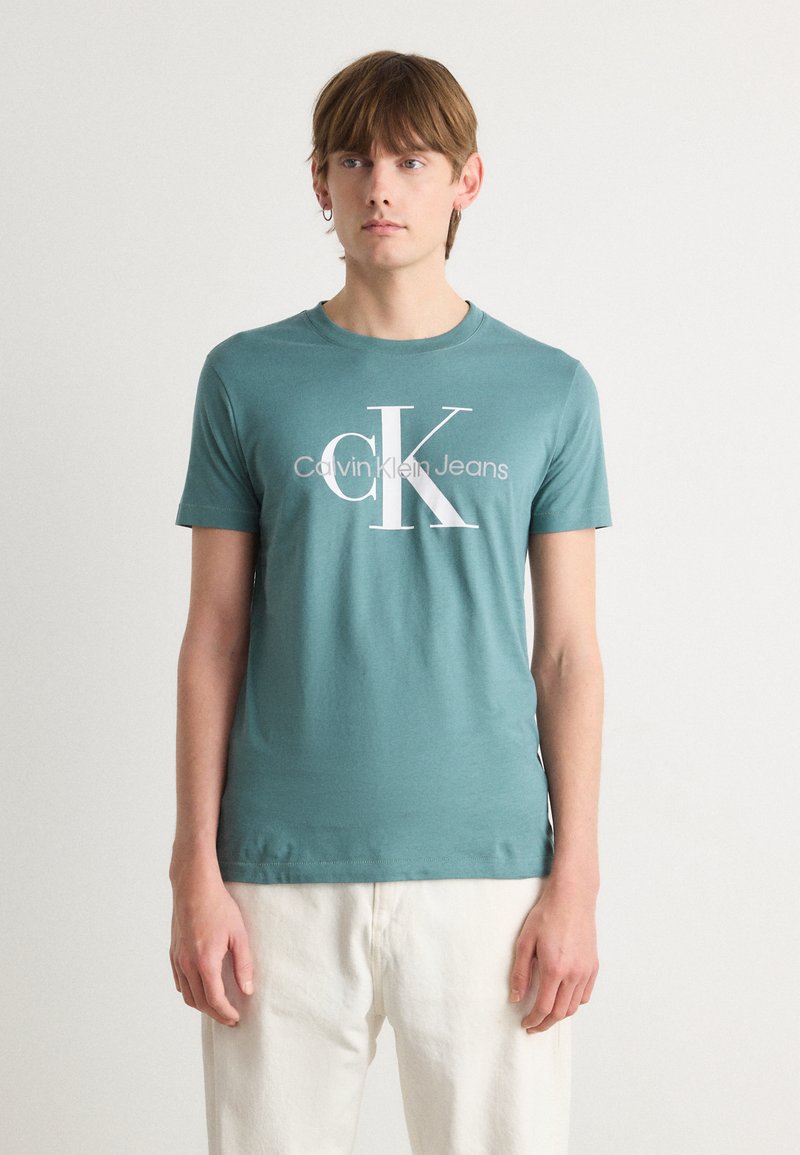 Žalias medvilnės marškinėliai su apvaliu iškirpimu ir trumpomis rankovėmis. Puošia didelė balta logotipo spauda, kuri sako "Calvin Klein Jeans."