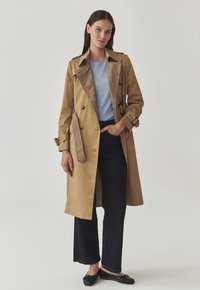 Beige trench coat with a double-breasted design, large front pockets, and contrasting brown plaid lining. Worn over a light blue top and dark jeans.