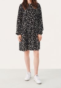 Abito nero con pois bianchi, maniche lunghe, scollatura rotonda, vita arricciata e gonna a strati. Abbinato a sneakers bianche.