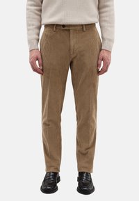 Hellbraune Cordhosen mit schmaler Passform, vertikalen Rippen, seitlichen Taschen und Knopfverschluss am Bund. Kombiniert mit schwarzen Schuhen.