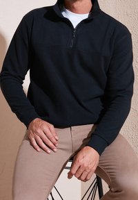 Pullover nero a mezza zip in un morbido tessuto con polsini e orlo a coste. Abbinato a pantaloni beige chiaro, seduto contro uno sfondo testurizzato.