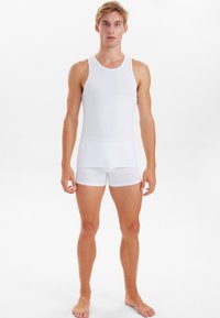 Weißes Baumwoll-Top ohne Ärmel und Boxershorts mit enganliegendem Design und glatter Textur, präsentiert an einem stehenden Modell vor einem neutralen Hintergrund.
