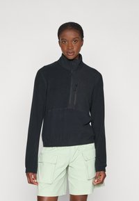 Schwarzer Fleece-Pullover mit hohem Kragen und halbem Reißverschluss; verfügt über eine Reißverschlusstasche auf der Brust und einen elastischen Bund. Getragen mit hellgrünen Cargo-Shorts.