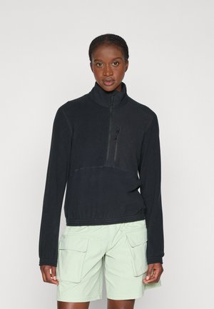 Svart fleecegenser med høy krage og halv glidelås foran; har en glidelåslomme på brystet og elastisk midje. Kombinert med lysegrønne cargo-shorts.