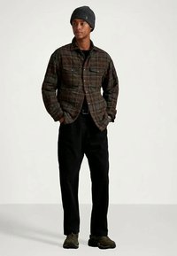 Young man wearing dark plaid jacket, black pants, gray knit beanie, and brown sneakers standing with hands in pockets.